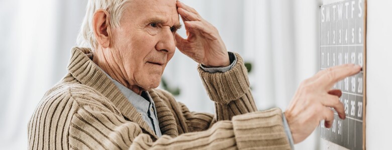 älterer Mann vor Kalenderblatt stehend mit Hand an Stirn schaut dieses verunsichert an